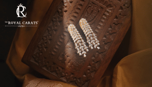 Pair of sparkling gold and diamond earrings resting on ornate wooden box with The Royal Carats branding.